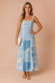 Vrouw draagt een lichtblauwe en witte maxi-jurk met bloemenpatchwork, dunne bandjes en kanten afwerking, en houdt een witte clutch vast tegen een warme perzikkleurige achtergrond.