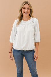Witte dames top met ronde hals en driekwartmouwen, gemaakt van soepele jersey stof. De bovenkant is voorzien van elegante plooien en een kleine split bij de halslijn. Elastische mouwboorden en een subtiel elastiek aan de onderzijde zorgen voor een comfortabele en luchtige pasvorm.